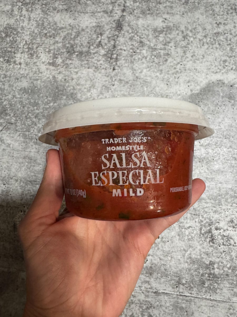 Hand holding Trader Joe's Salsa Especial Mild in a clear container against a textured gray backgroun.