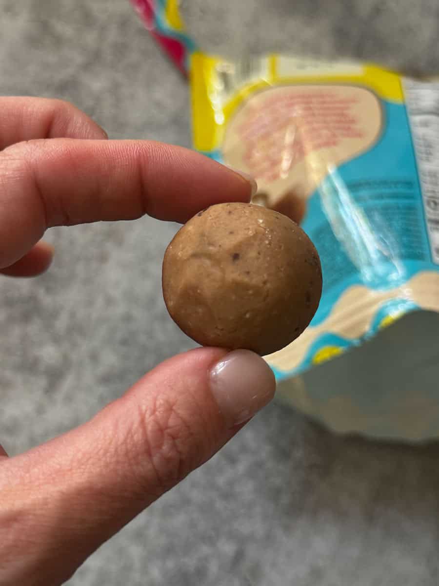 Hand holding a small snack ball with a bag of snack in the background.