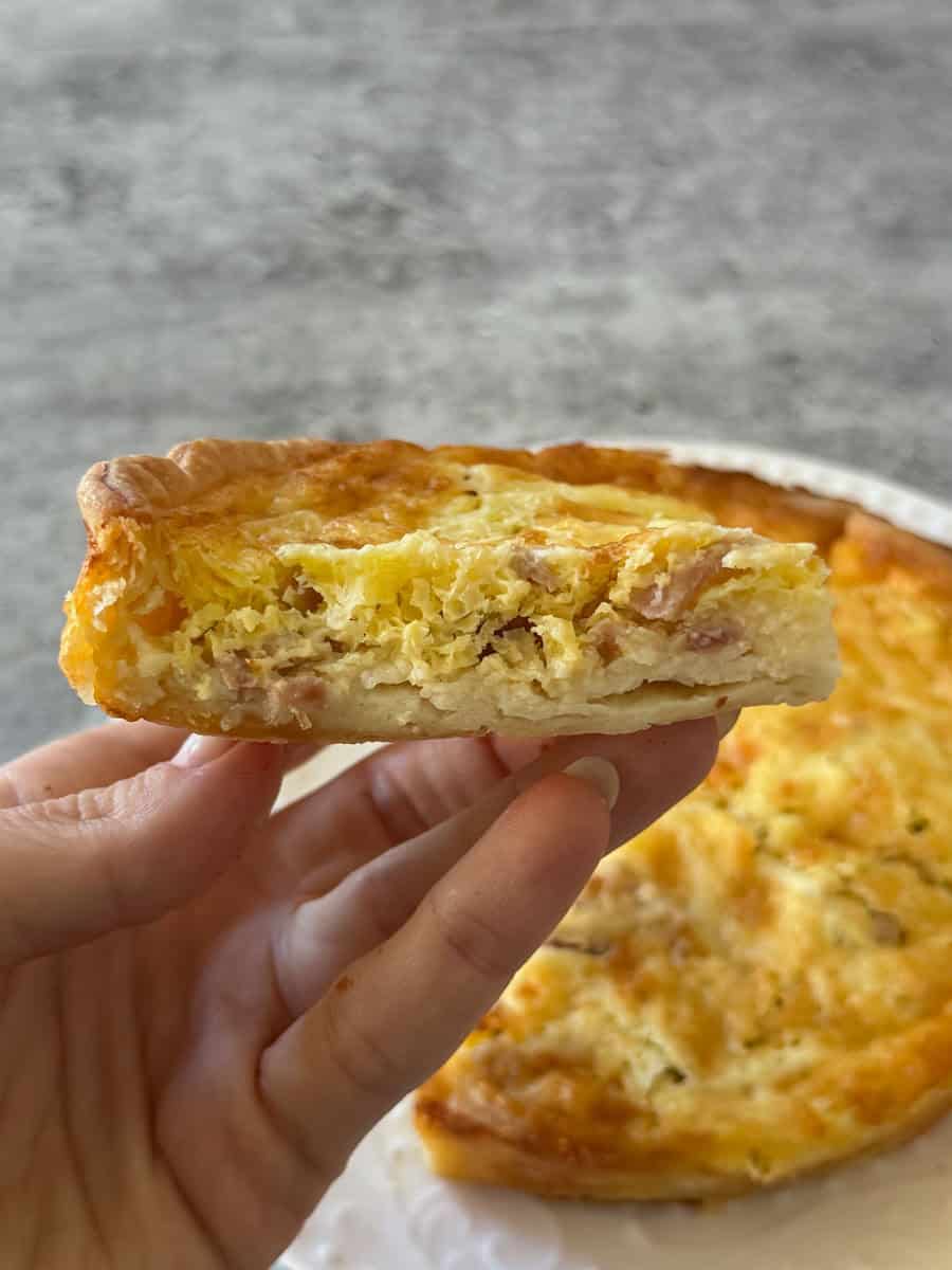 Hand holding a slice of cheese and ham quiche with a golden crust, fresh from the pie.